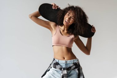 Beyaz duvarın üzerinde kaykayla poz verirken gülen Afrikalı genç bir kadın.