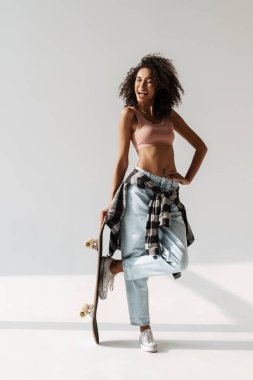 Beyaz duvarın üzerinde kaykayla poz verirken gülen Afrikalı genç bir kadın.