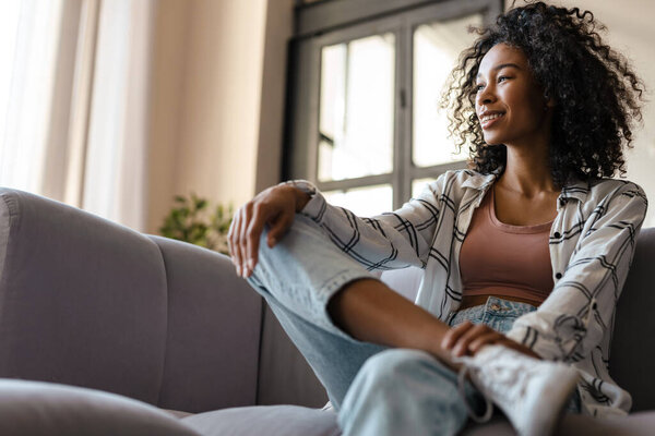 Улыбающаяся молодая африканская женщина в повседневной одежде держит мобильный телефон, сидя дома на диване