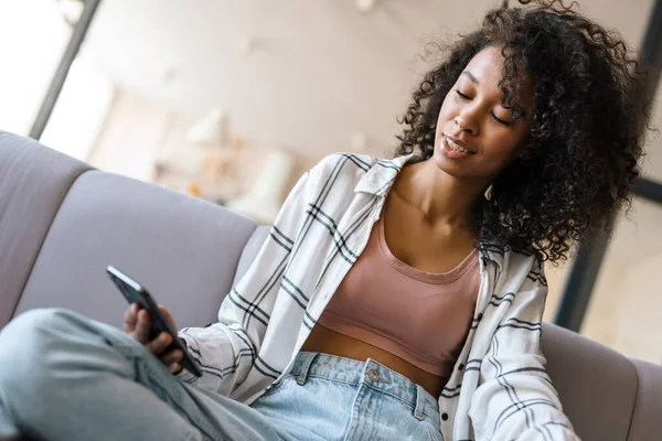 stock image Smiling young african woman in casual wear holding mobile phone while sitting on a couch at home