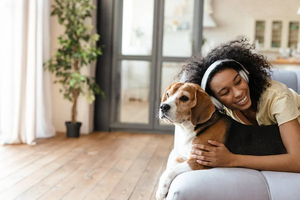 Happy young african woman in headphones relaxing on a couch with her pet dog at home, cuddling