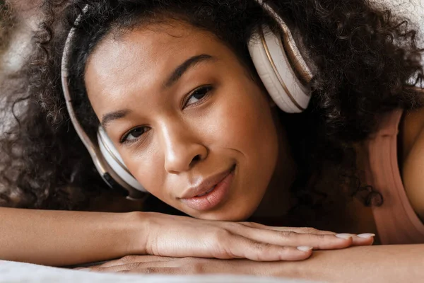 Smiling mid aged african woman listening to music with headphones while relaxing on bed at home, close up