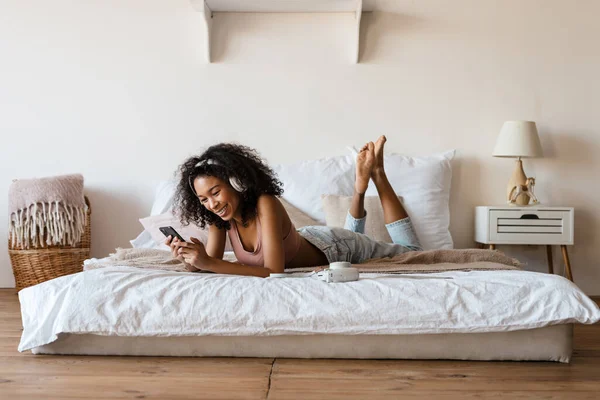 Smiling mid aged african woman listening to music with headphones while relaxing on bed at home, looking at mobile phone in her hands