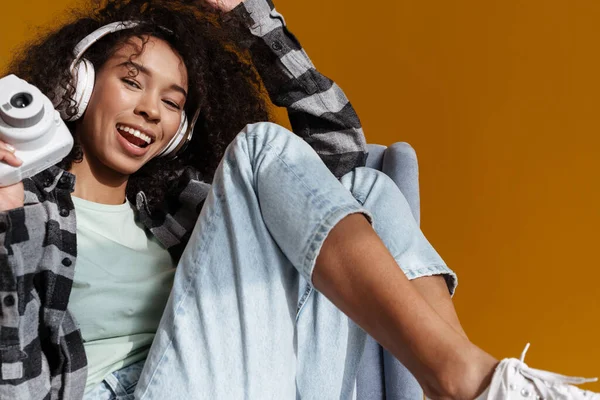 Young african woman in headphones posing with camera isolated over brown wall