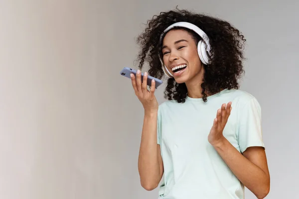 Young african woman in headphones smiling while using mobile phone isolated over white wall