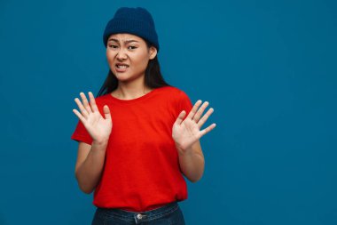 Şapkalı Asyalı genç kız mavi arka planda izole edilmiş dur hareketi gösteriyor.