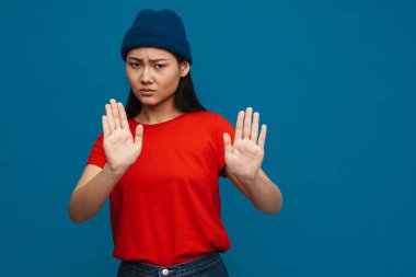 Şapkalı Asyalı genç kız mavi arka planda izole edilmiş dur hareketi gösteriyor.