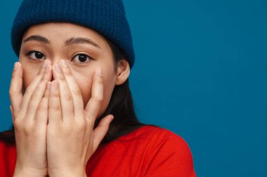 Şok olmuş, sıradan giyinmiş, mavi duvar arkasında duran, yüzünü ellerle kaplayan Asyalı genç bir kadın.