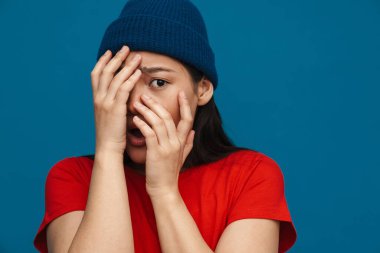 Şok olmuş, sıradan giyinmiş, mavi duvar arkasında duran, elleri ile yüzünü kapatan Asyalı genç bir kadın.