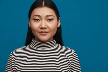 Mavi duvarın arkasında dikilip kameraya bakan sıradan giysili genç Asyalı kadın.