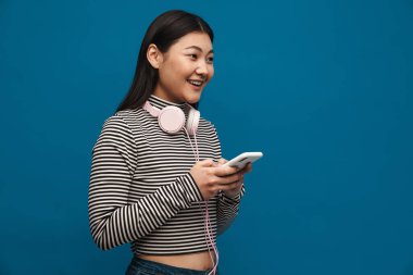 Mavi duvar arkasında elinde cep telefonu olan, sıradan giyinmiş mutlu Asyalı genç bir kadın.
