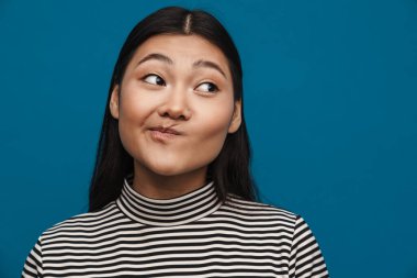 Kafası karışmış Asyalı genç kız mavi arka planda yalnız poz veriyor ve bir kenara bakıyor.