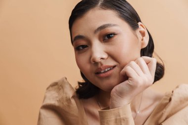 Genç Asyalı esmer kadın poz veriyor ve bej arka planda izole edilmiş kameraya bakıyor.