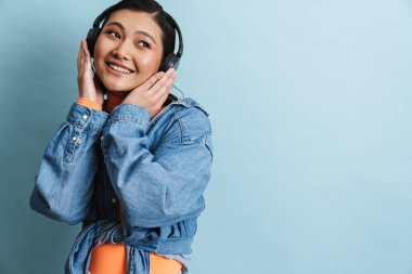 Kot ceketli mutlu Asyalı genç esmer kadın mavi arka planda duruyor kulaklıkla müzik dinliyor.