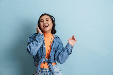 Kot ceketli mutlu Asyalı genç esmer kadın mavi arka planda duruyor kulaklıkla müzik dinliyor.