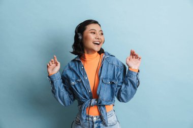 Kot ceketli mutlu Asyalı genç esmer kadın mavi arka planda duruyor kulaklıkla müzik dinliyor.