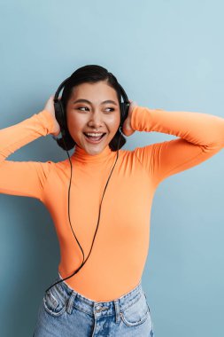 Mavi arka planda izole edilmiş kulaklıklarla müzik dinlerken gülümseyen Asyalı genç bir kadın.