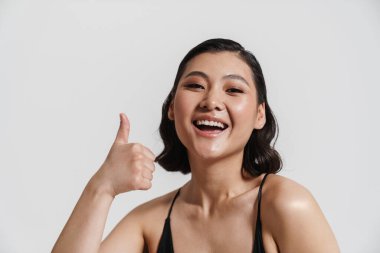 Mayo giymiş esmer bir kadın gülümsüyor ve beyaz arka planda baş parmağını gösteriyor.