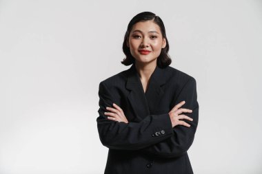 Ceketli genç beyaz Asyalı kadın beyaz duvar arkasında durup kamera kollarına katlanmış bir şekilde bakıyor.