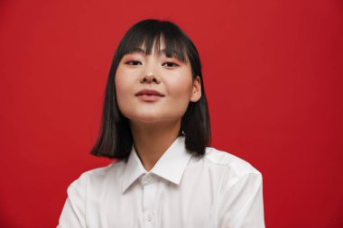 Genç Asyalı esmer kadın kırmızı arka planda yalnız poz veriyor ve kameraya bakıyor.