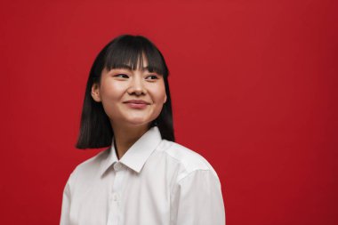 Genç Asyalı esmer kadın gülümsüyor ve kırmızı arka planda soyutlanmış bir şekilde yan tarafa bakıyor.