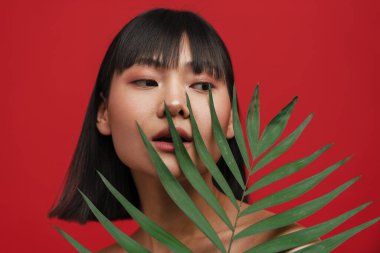 Asyalı üstsüz kadın kırmızı arka planda yeşil yapraklı poz verirken yana bakıyor.