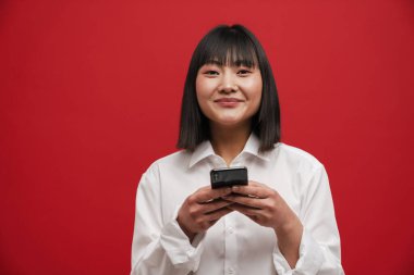 Kırmızı arka planda cep telefonu kullanırken gülümseyen Asyalı genç bir kadın.