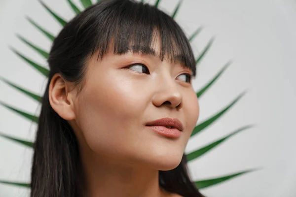 Young asian woman looking aside while posing with green leaf isolated over white background