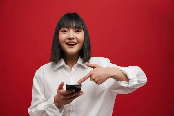 年轻的亚洲女人一边微笑着 一边用手指指指着红色背景下孤立的手机 — 图库照片