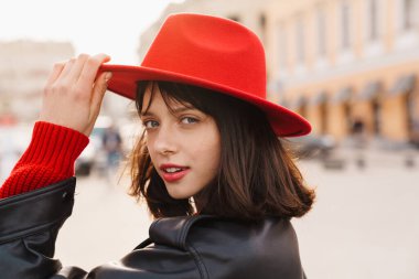 Gülümseyen, şık, esmer, kısa saçlı, şapkalı, deri ceketli beyaz bir kadının bir sokakta durup kameraya baktığı fotoğrafı.