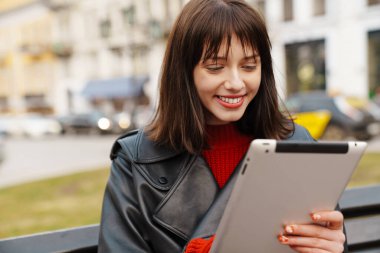 Elinde tablet olan deri ceketli gülümseyen beyaz bir kadın sokakta bir bankta oturuyor.