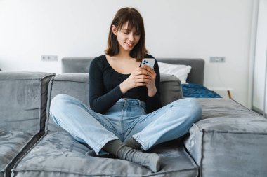 Evdeki koltukta otururken cep telefonu kullanan gülümseyen genç kadın.