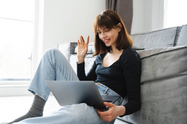 Evde dizüstü bilgisayarı olan gülümseyen beyaz bir kadın, bir video görüşmesinde, el sallıyor.