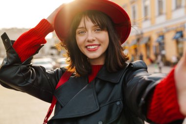 Gülümseyen, şık, esmer, kısa saçlı, şapkalı, deri ceketli beyaz bir kadının fotoğrafını çek.