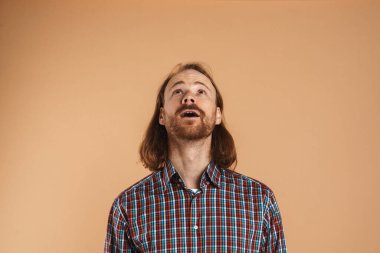 Kareli gömlek giyen sakallı kızıl adam bej arka planda yalnız poz veriyor ve yukarı bakıyor.