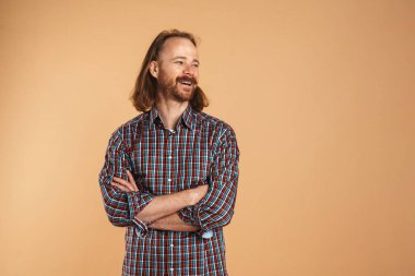 Kareli gömlek giyen sakallı kızıl adam gülümsüyor ve bej arka planda soyutlanmış bir kenara bakıyor.