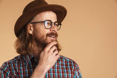 Gözlüklü, şapkalı, gülümseyen kızıl adam bej arka planda izole edilmiş bir yere bakıyor.