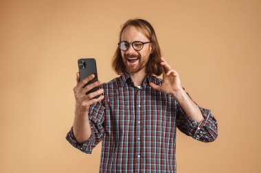Gülümseyen kızıl adam, bej arka planda izole edilmiş cep telefonuyla selfie çekerken işaret veriyor.
