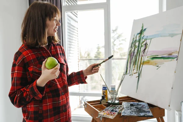 キャンバスの中に絵筆を持っている若い笑顔の白い女性画家は アップルを食べ — ストック写真