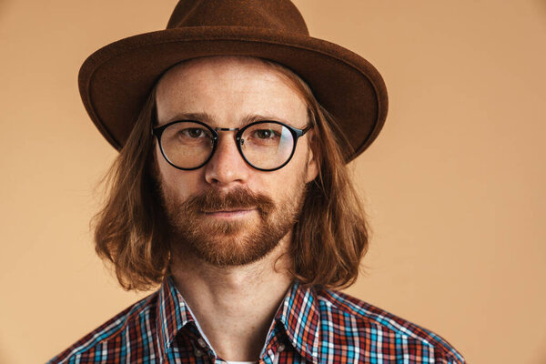 Bearded ginger man wearing eyeglasses and hat looking at camera isolated over beige background
