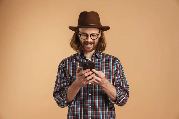 在米色背景下 用孤立的手机微笑着戴着眼镜和帽子的姜人 — 图库照片
