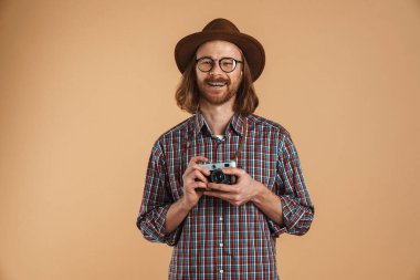 Gözlüklü ve şapkalı gülen kızıl adam bej arka planda izole edilmiş retro kamerayla poz veriyor.