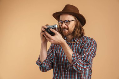 Tıraşsız kızıl adam gözlük takıyor ve şapka takıyor. Bej arka planda izole edilmiş retro kamera kullanıyor.