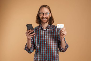 Gülümseyen kızıl adam, cep telefonu ve kredi kartıyla bej arka planda poz veriyor.