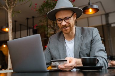 Beyaz sakallı adam dışarıda kafede otururken dizüstü bilgisayar ve kredi kartı kullanıyor.