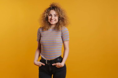 Sarı duvarın arkasında dikilmiş kameraya bakan, günlük kıyafetleri olan, mutlu, genç, sarışın bir kadın.