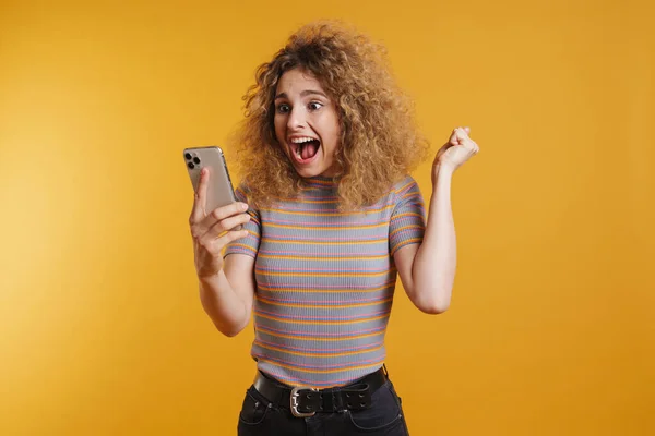 幸せな笑顔若いですブロンド女性とともにFizzy毛でカジュアルな服に立って上の黄色の壁の背景 携帯電話を保持し 祝います — ストック写真