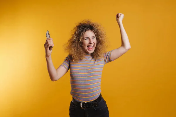 年轻的金发女子 穿着休闲装 笑容满面地站在黄墙的后面 拿着手机庆祝 — 图库照片