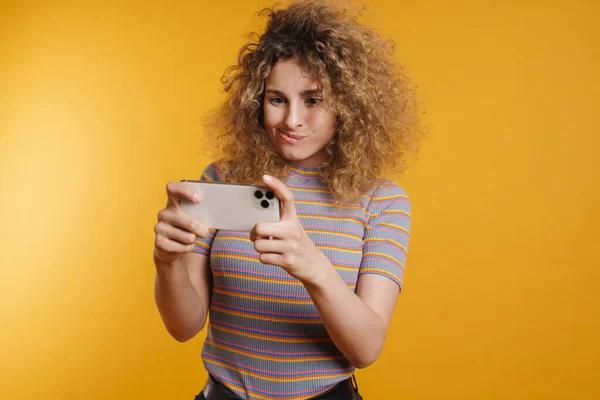 若いですブロンド女性とともに固定毛でカジュアルな服の上に立っている黄色の壁の背景 携帯電話でゲームをプレイ — ストック写真