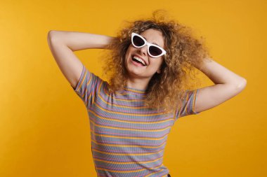 Şen şakrak saçlı, güneş gözlüğü takan, sarı duvarın arkasında dans eden, kutlama yapan mutlu bir genç kadın.